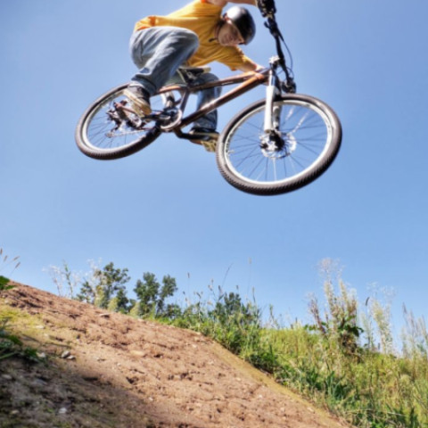 Chlapec na bicykli skáče cez prekážku na modrom pozadí s oblohou a zeleňou, ilustrujúci bikepark pre mladých v Senici.