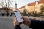 Ruky držiace mobil s rozmazanou diskusiou, v pozadí námestie malého mesta v jesennom svetle