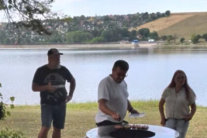 Traja ľudia stoja pri veľkom vonkajšom ohnisku pri vode.