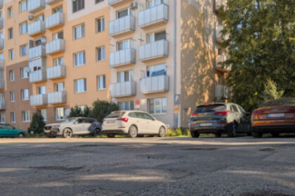 Poškodený asfalt na ulici pred bytovým domom so zaparkovanými autami.
