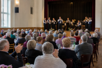 Seniori sediaci v kultúrnom dome počas hudobného vystúpenia dychovej hudby.
