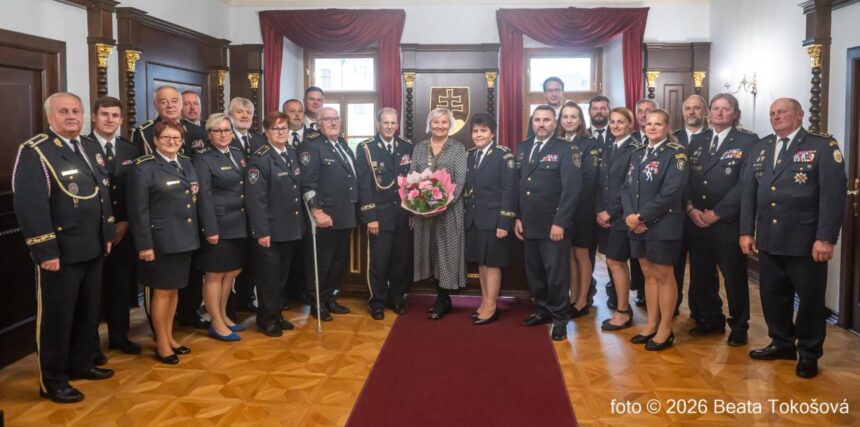 Skupina ľudí v hasičských uniformách v interiéri, jedna osoba drží kyticu.