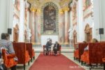 Interiér jezuitského kostola v Skalici počas koncertu, na pódiu sedia dve osoby a v hľadisku sú diváci.
